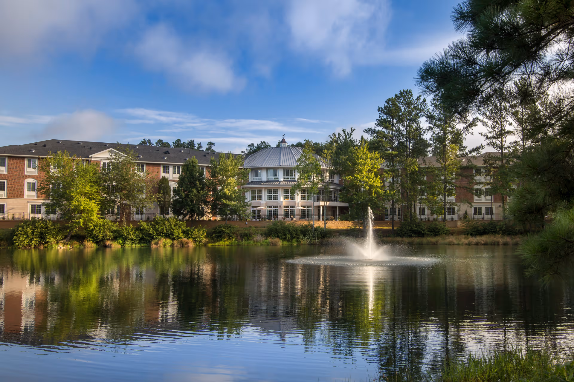 A scenic view of The Georgian Lakeside facility showing a large pond with a fountain in the center, surrounded by trees and greenery. The multi-story building with a mix of brick and light-colored siding is visible in the background under a partly cloudy blue sky.