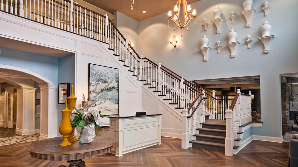 Elegant lobby with a round table topped by vases and flowers, a reception desk, and a grand staircase leading to an upper gallery.