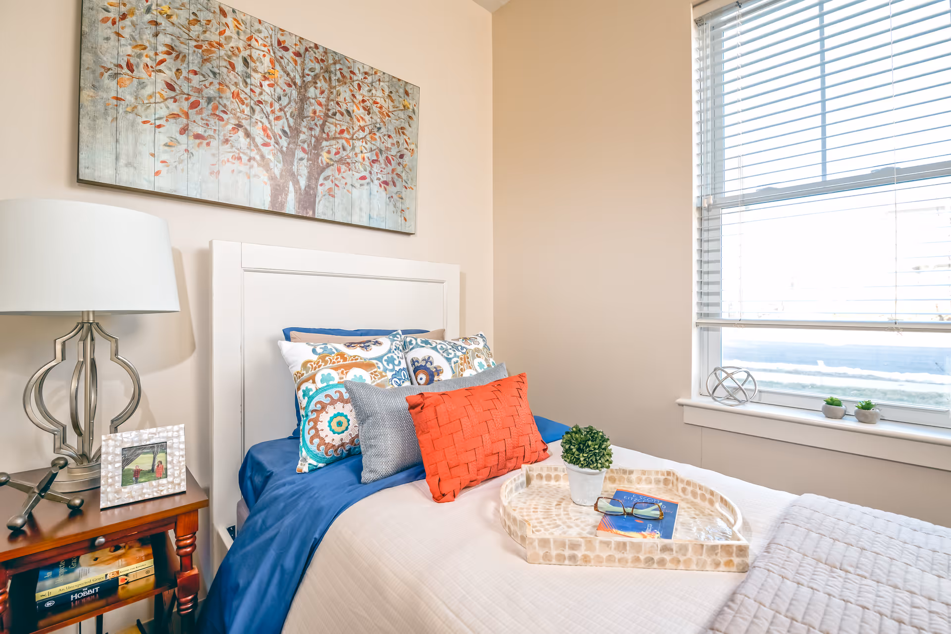 A cozy bedroom with a white headboard bed dressed in blue and white bedding, colorful decorative pillows, and a beige quilt. A wooden nightstand with a modern lamp, a framed photo, and books is beside the bed. Above the bed hangs a large painting of a tree with autumn leaves. A window with white blinds lets in natural light, and a tray with a small potted plant, glasses, and a book rests on the bed.