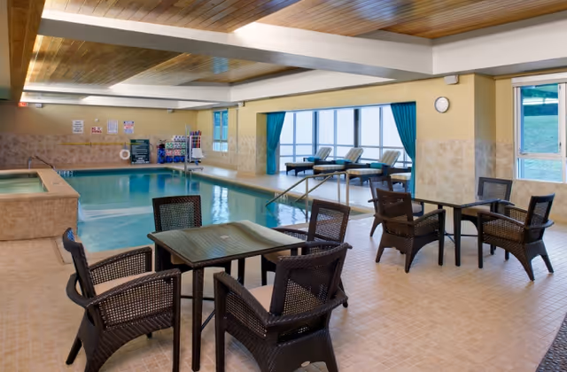 Indoor swimming pool area with several wicker chairs and tables arranged on the tiled floor. The pool has handrails and a hot tub section on the left side. Large windows with blue curtains let in natural light, and there are lounge chairs near the windows.
