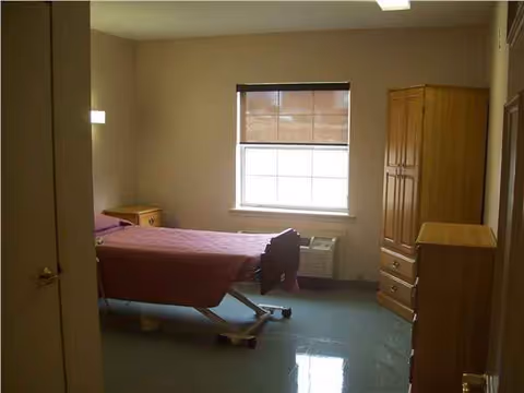Adjustable hospital-style bed in a senior facility bedroom with dresser, wardrobe, and window.