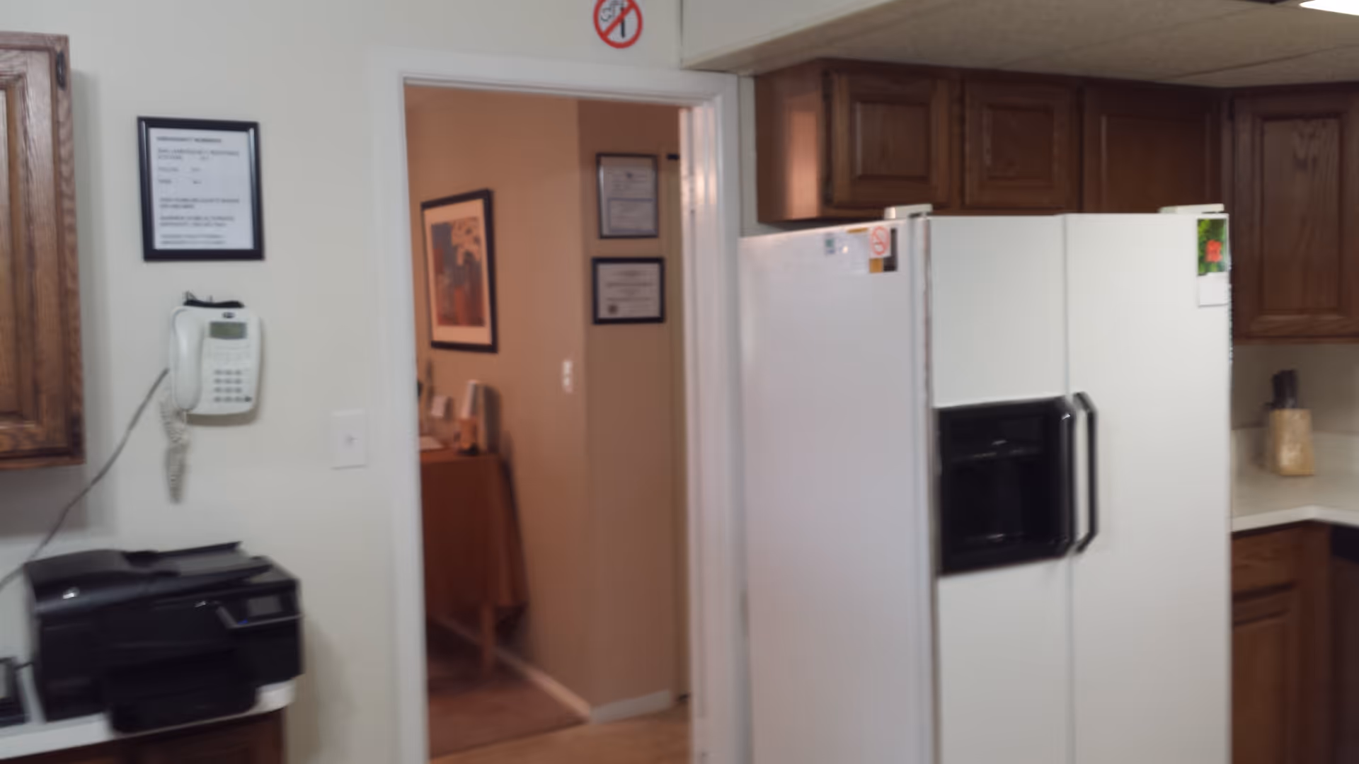 Interior view of a kitchen area showing a white refrigerator with a water and ice dispenser, wooden cabinets, a wall-mounted telephone, a printer on a counter, and a doorway leading to another room with framed certificates and artwork on the walls.
