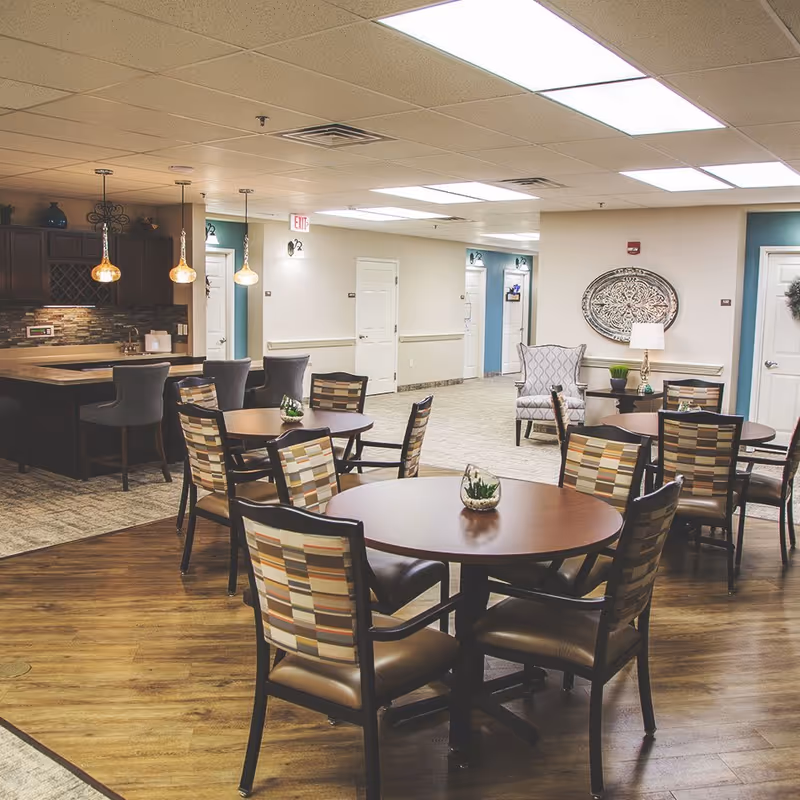 Interior view of a senior living facility common area featuring round tables with patterned chairs, a small kitchen area with bar stools and pendant lights, and a hallway with several closed doors. The space is well-lit with ceiling lights and decorated with wall art and plants.