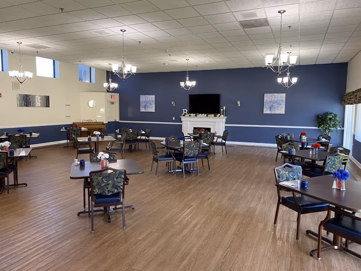 A spacious dining room with several tables and chairs arranged neatly. Each table has a small flower arrangement. The room features wood flooring, blue and white walls, multiple chandeliers hanging from the ceiling, a fireplace with a TV mounted above it, and large windows allowing natural light to enter.