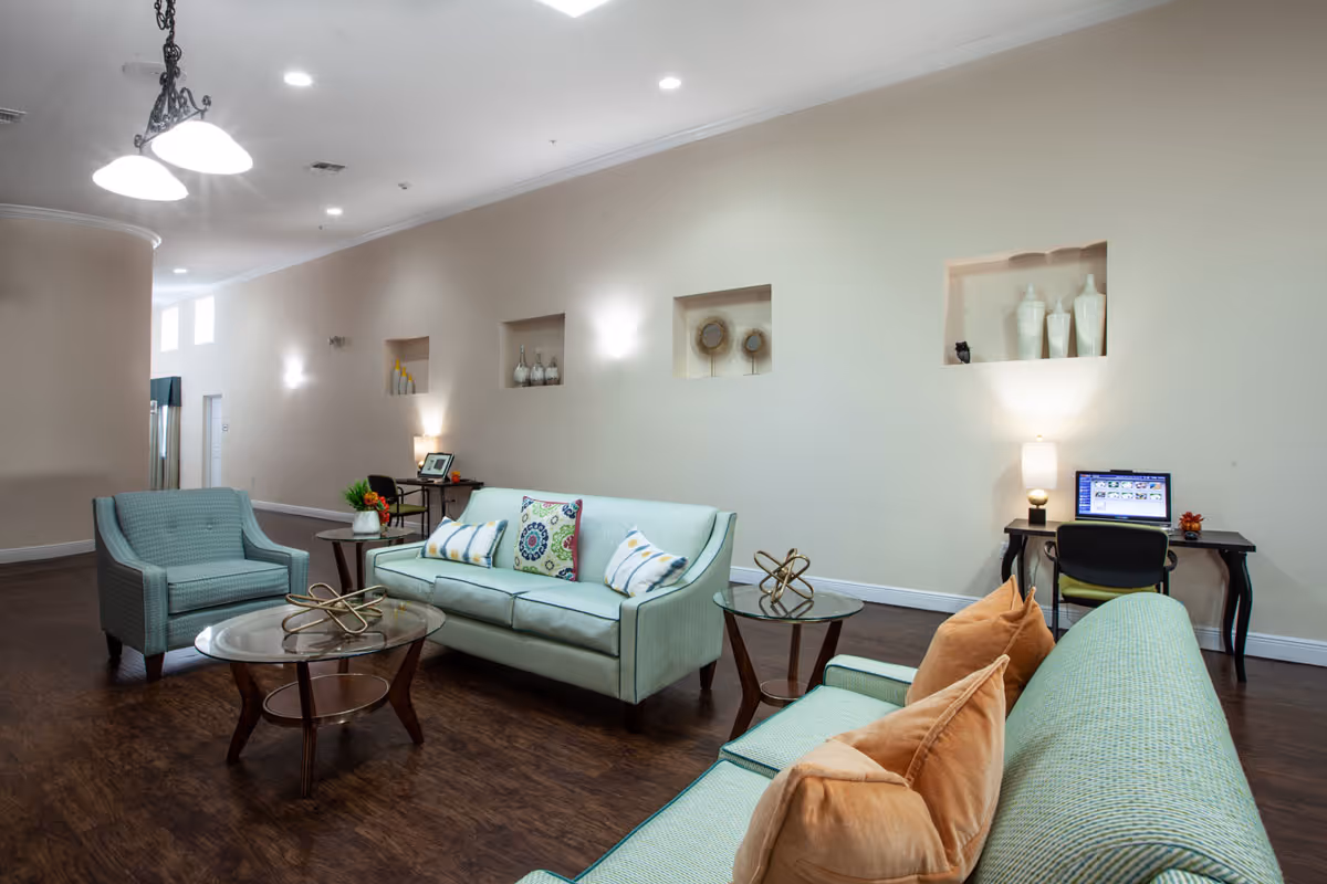 A spacious and well-lit living room area with light green upholstered sofas and armchairs arranged around glass-top wooden coffee and side tables. The walls are beige with built-in shelves displaying decorative vases and art pieces. There are two small desks with chairs and lamps along the wall, one with a laptop on it. The floor is dark wood, and ceiling lights illuminate the room.