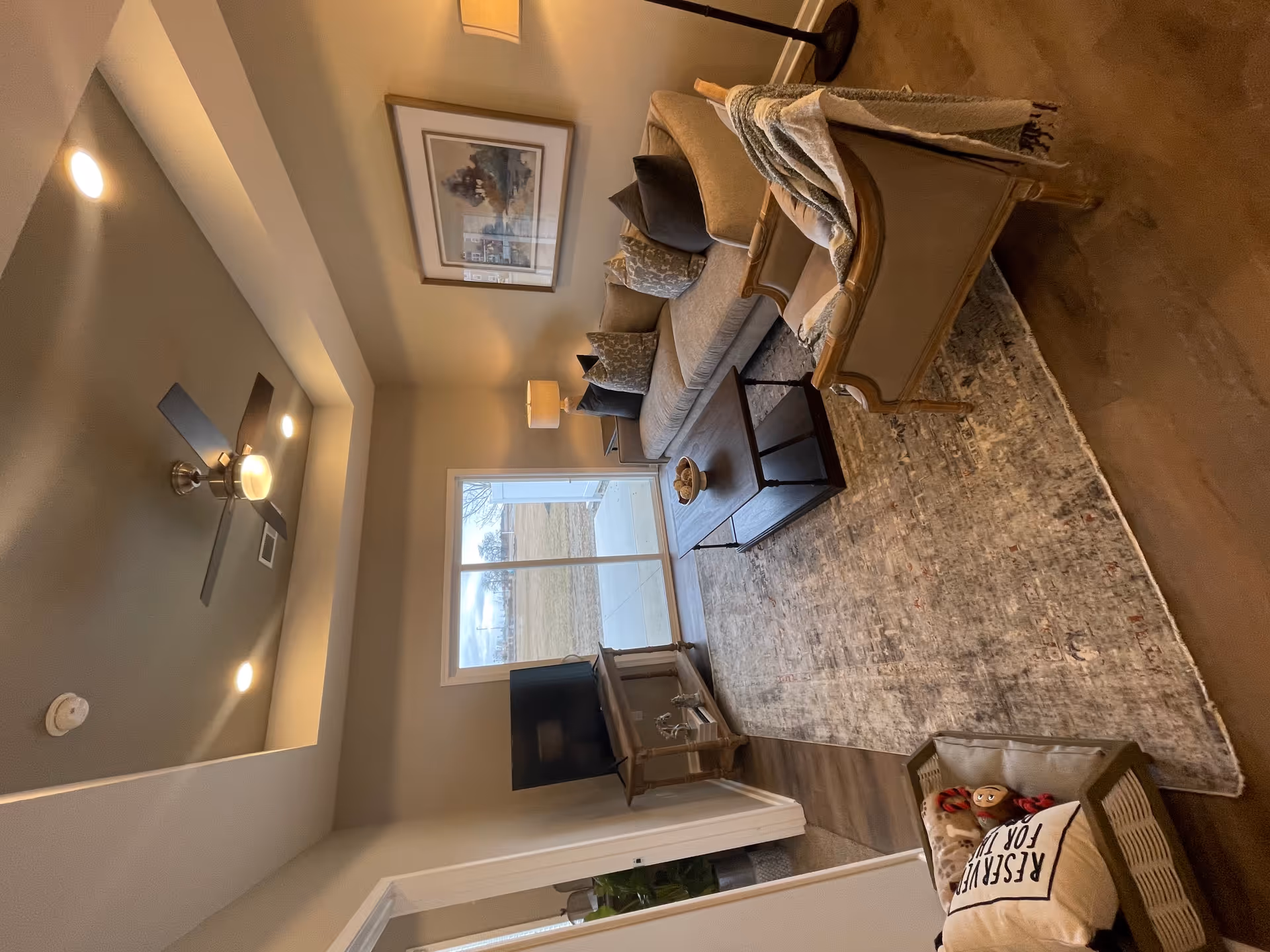 A cozy living room with a beige sofa adorned with multiple cushions, a wooden coffee table with a decorative bowl, an armchair with a throw blanket, a TV on a stand, a large window showing an outdoor view, a ceiling fan with lights, a framed picture on the wall, and a pet bed with a pillow and toy on the floor.