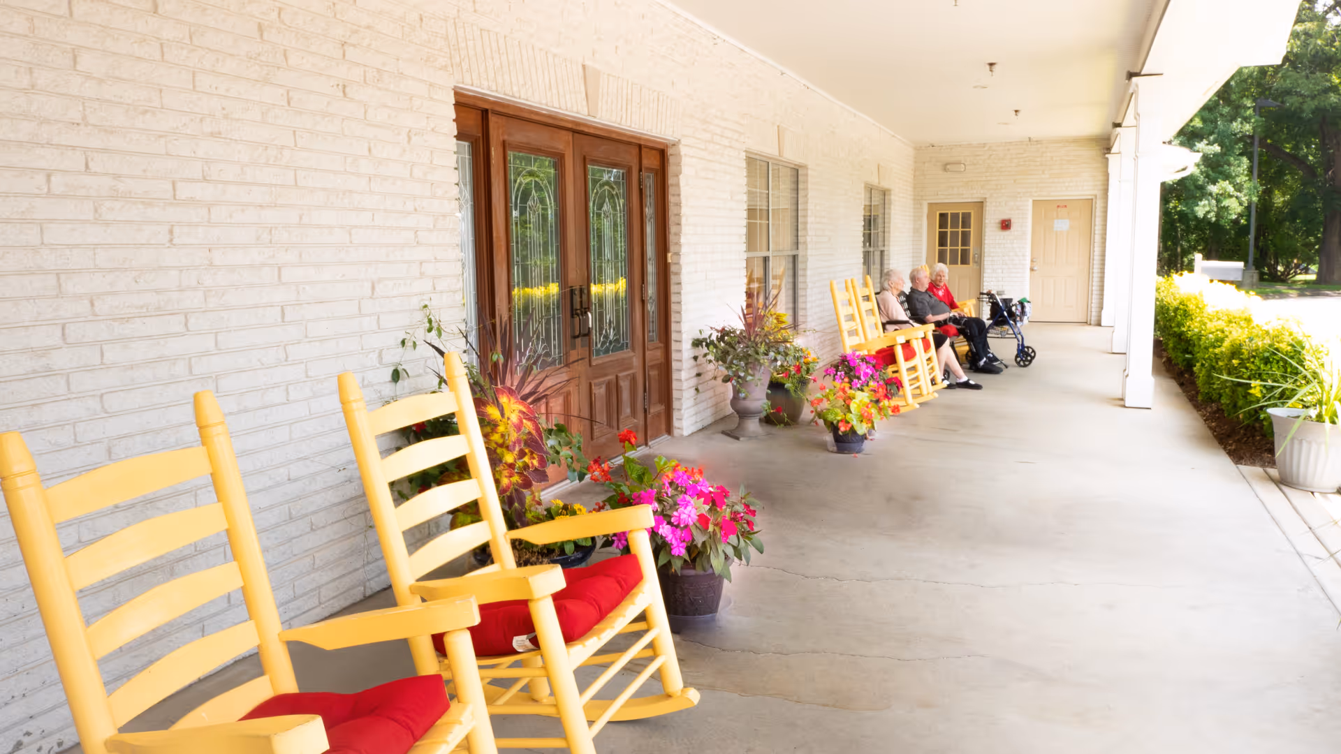 A covered outdoor porch area with yellow rocking chairs that have red cushions, several potted flowering plants, and three elderly people sitting and chatting near the far end. The porch is attached to a white brick building with wooden double doors and windows.