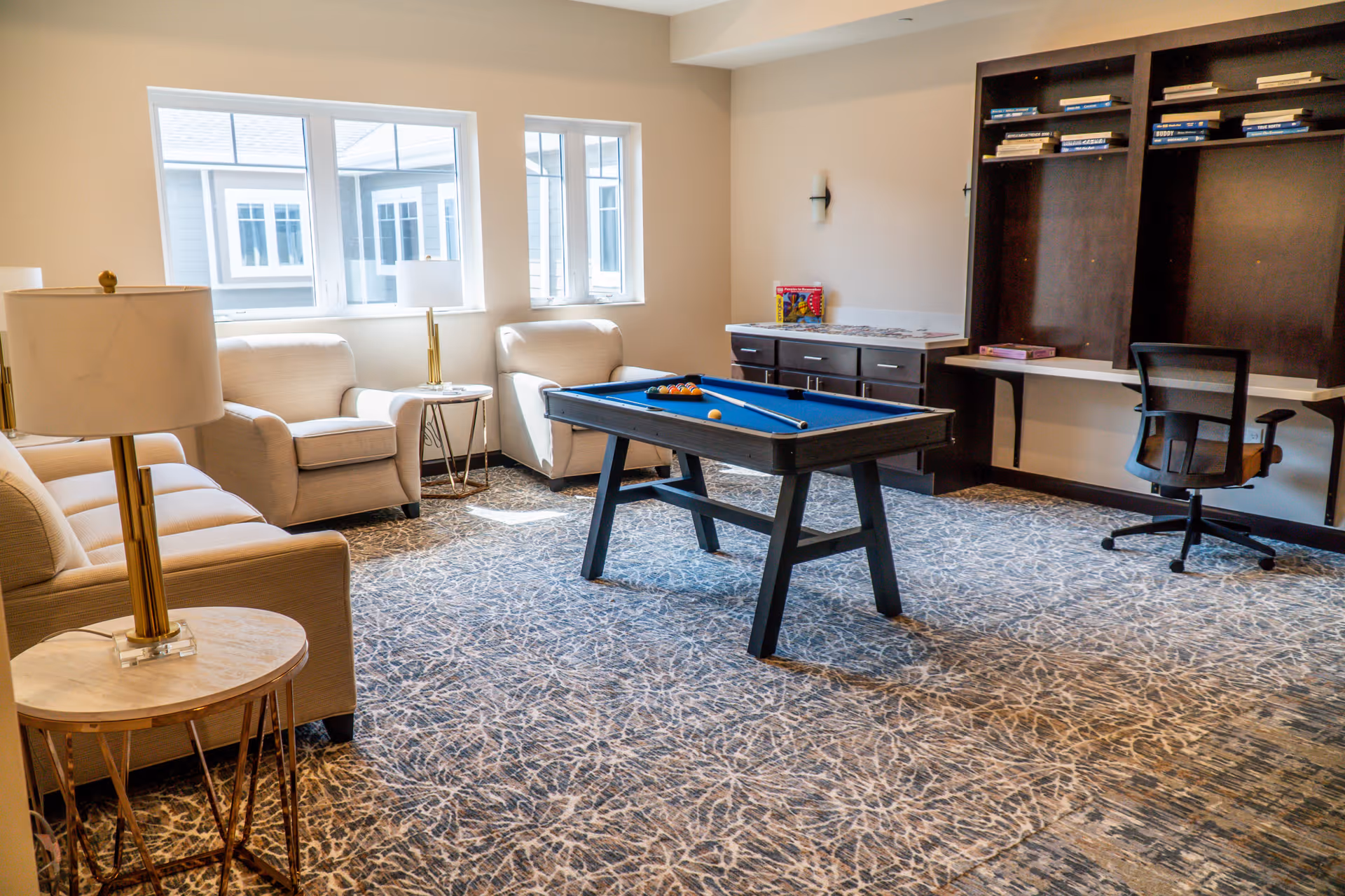 Bright communal lounge with a small pool table centered, upholstered chairs and sofas, side tables, lamps, and a desk with shelving.