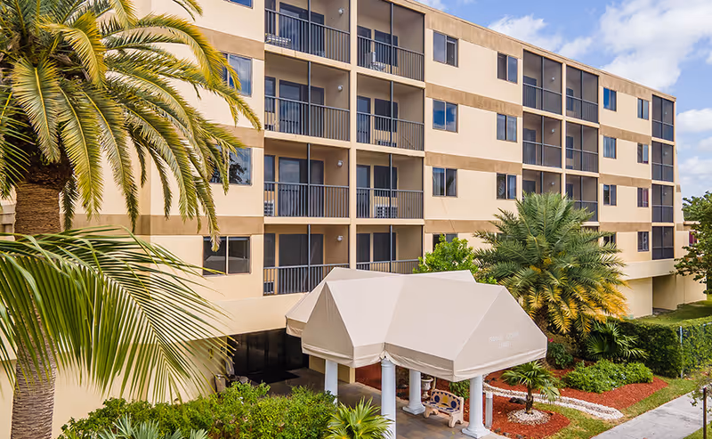 Exterior view of a multi-story residential building with balconies, surrounded by palm trees and landscaped greenery. A covered entrance canopy is visible at the front with the name Grand Court Lakes on it.