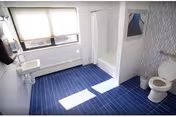 Bright accessible bathroom with a blue-tiled floor, white sink, toilet, and a tub/shower next to a large window.