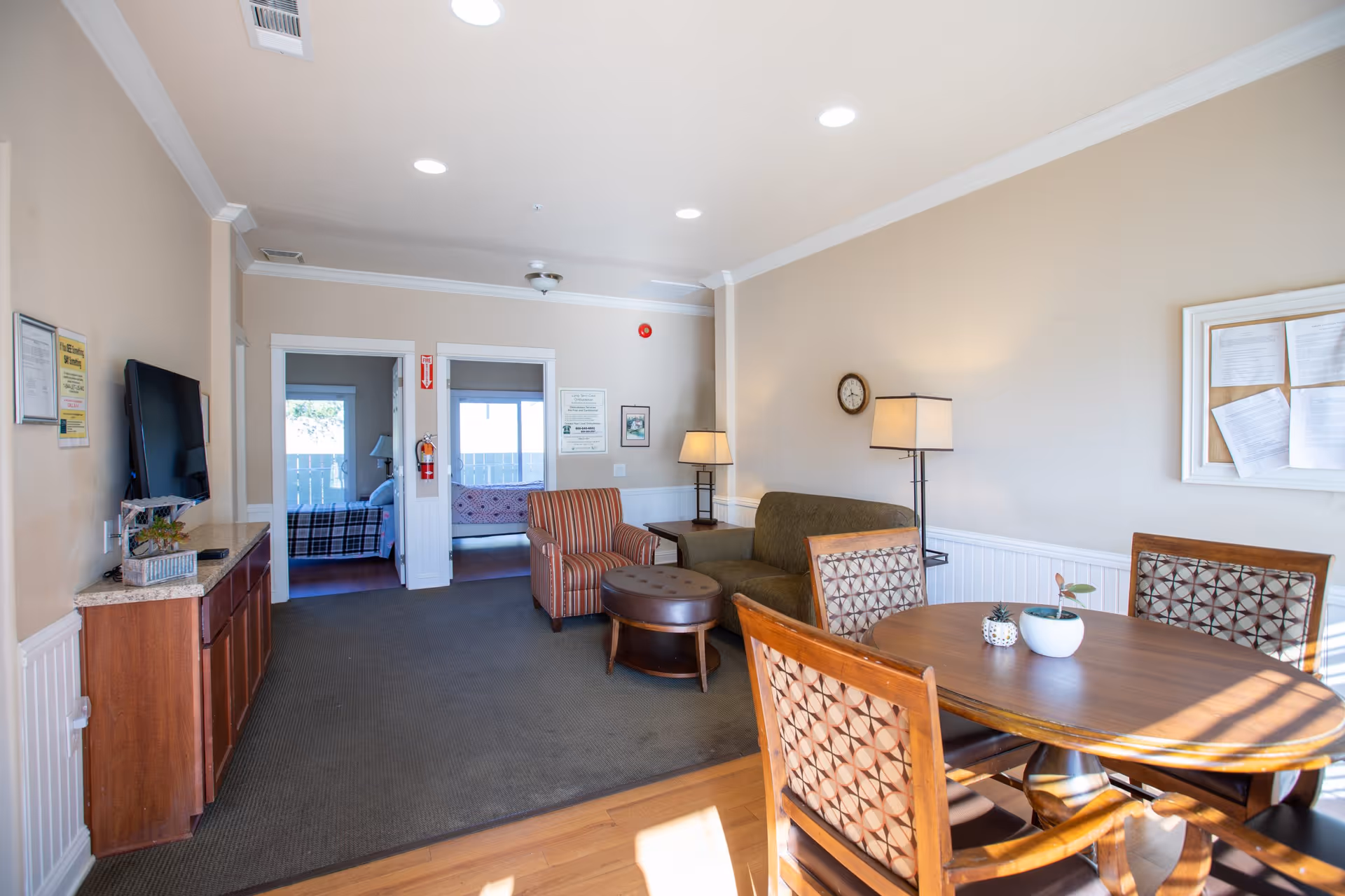 A well-lit senior living common area with a round wooden table and four patterned chairs in the foreground. To the right, there is a green sofa, a striped armchair, a round ottoman, and two lamps. A flat-screen TV is mounted on the left wall above a wooden cabinet. Two doorways in the back lead to bedrooms with beds visible. The walls are beige with white trim, and there is a bulletin board with papers on the right wall.