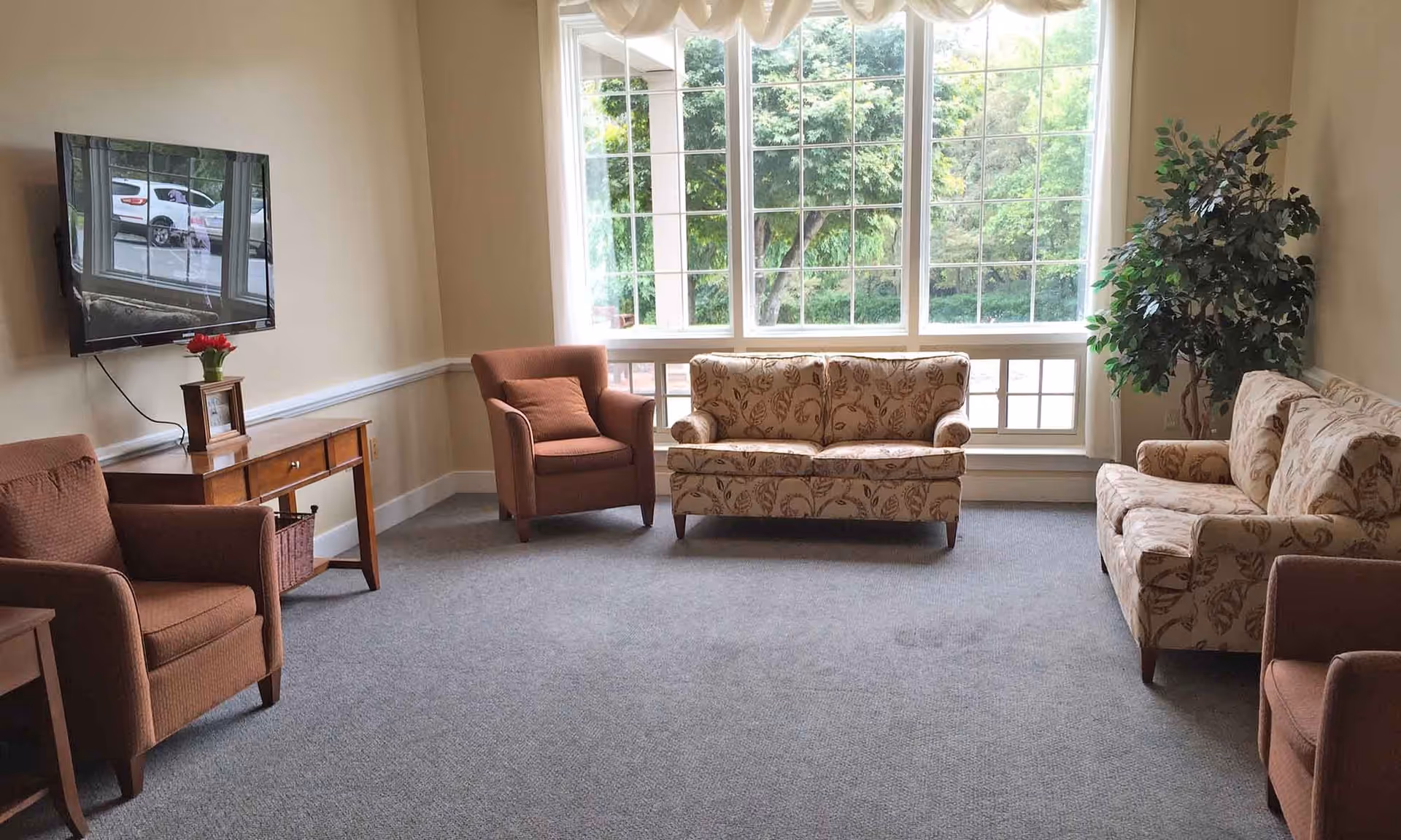 A cozy living room with a large window showing green trees outside. The room has two patterned sofas, two brown armchairs, a wooden side table with a small plant and a framed photo, and a wall-mounted flat-screen TV.