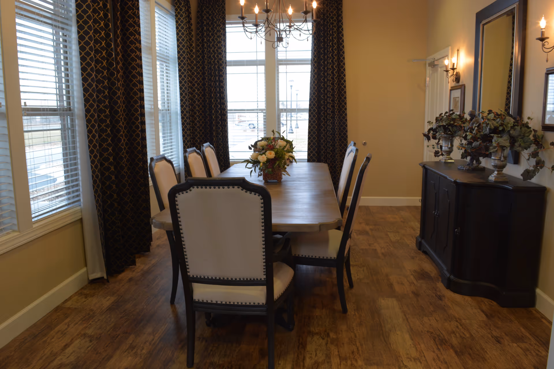 Formal dining room with a wooden table surrounded by upholstered chairs, a chandelier overhead, large windows with patterned curtains, and a sideboard with decorative plants.