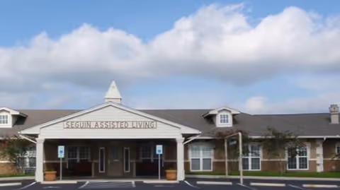 Exterior front view of Seguin Assisted Living facility showing a single-story building with a covered entrance, multiple windows, and a sign displaying the facility name. The sky is partly cloudy.