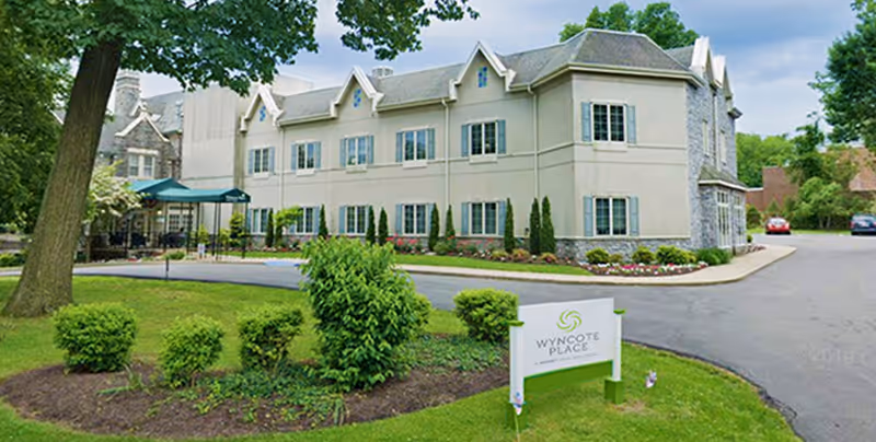 Exterior view of Wyncote Place, a two-story building with multiple windows, surrounded by green trees, shrubs, and a well-maintained lawn. A sign with the facility name is visible in the foreground near the driveway.