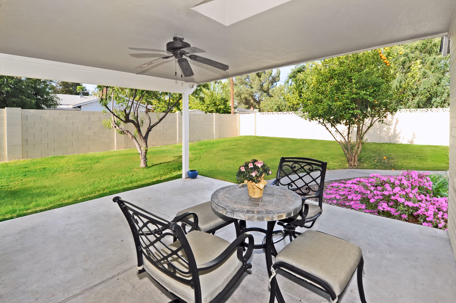 Covered outdoor patio area with a round stone table and four cushioned chairs. A small potted plant with pink flowers is on the table. The patio overlooks a green lawn with trees, a flower bed with pink flowers, and a white fence in the background.