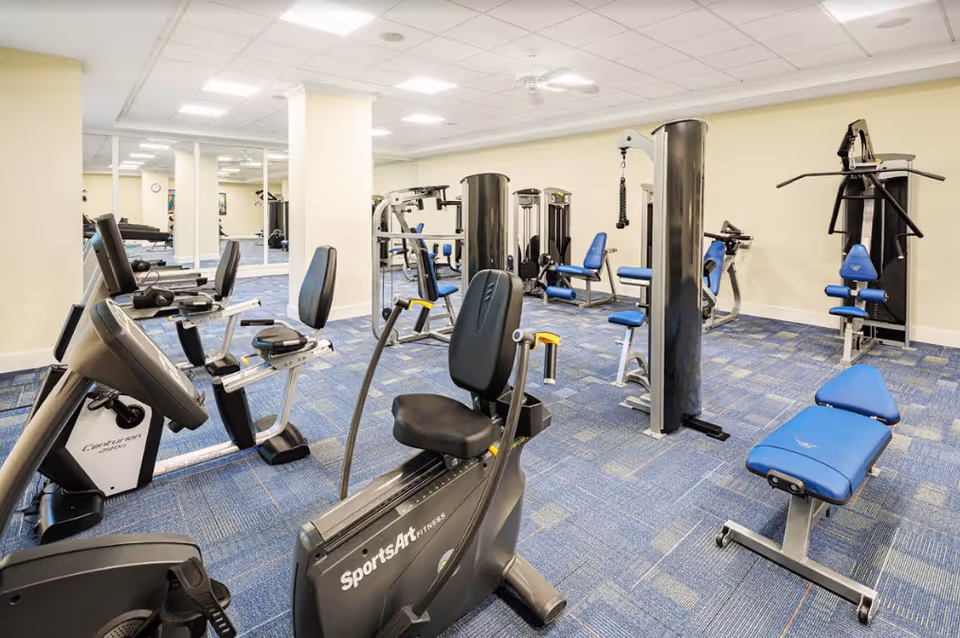 A fitness room in a senior living facility featuring various exercise equipment including stationary bikes, weight machines, and benches. The room has blue carpet flooring, light-colored walls, and a ceiling with recessed lighting and a ceiling fan. Mirrors line one wall, reflecting the equipment and space.