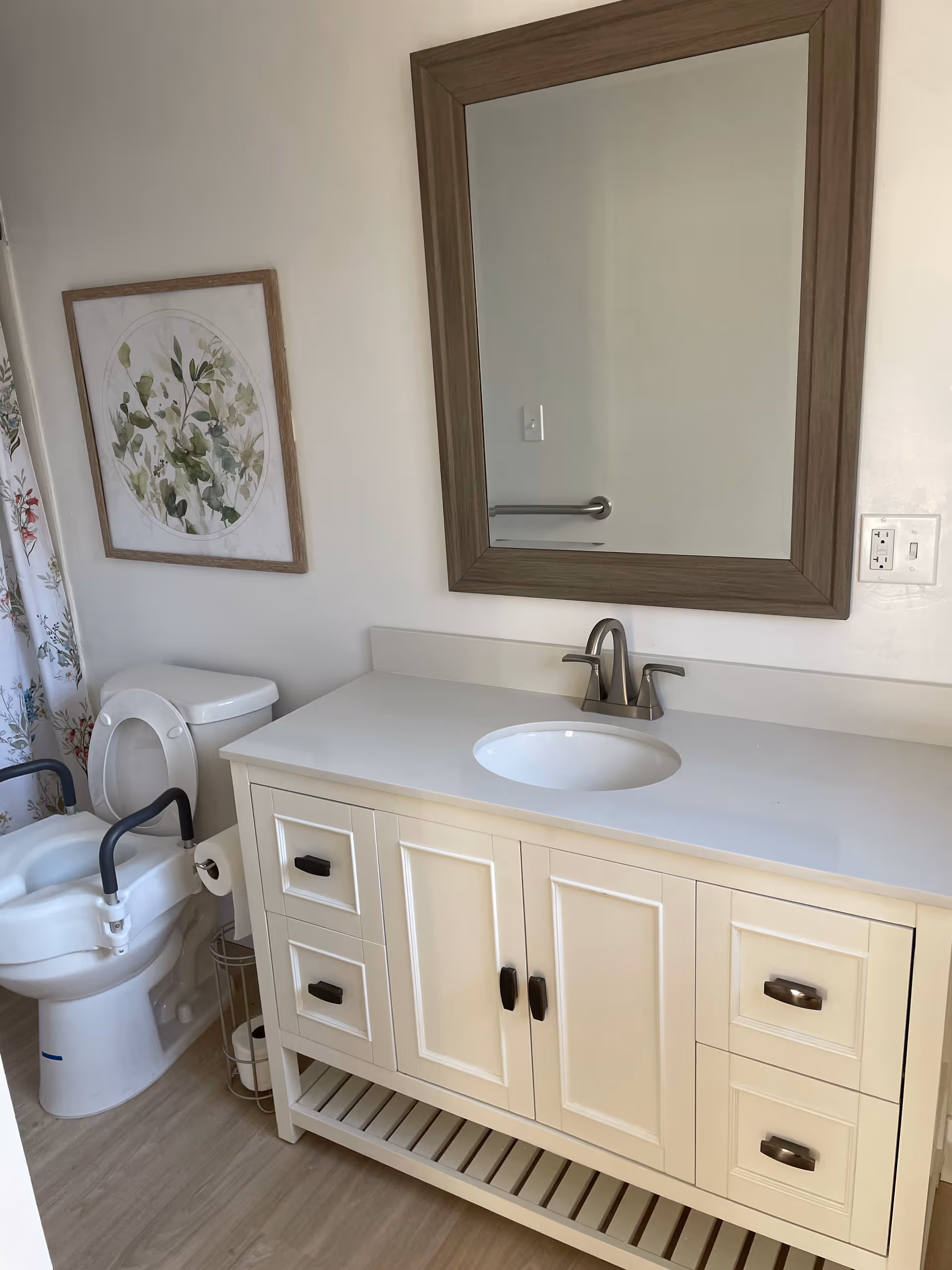 A clean bathroom featuring a white vanity with a sink and silver faucet, a large rectangular mirror with a wooden frame above the vanity, a toilet with a raised seat and armrests, a floral shower curtain, and a framed botanical artwork on the wall.