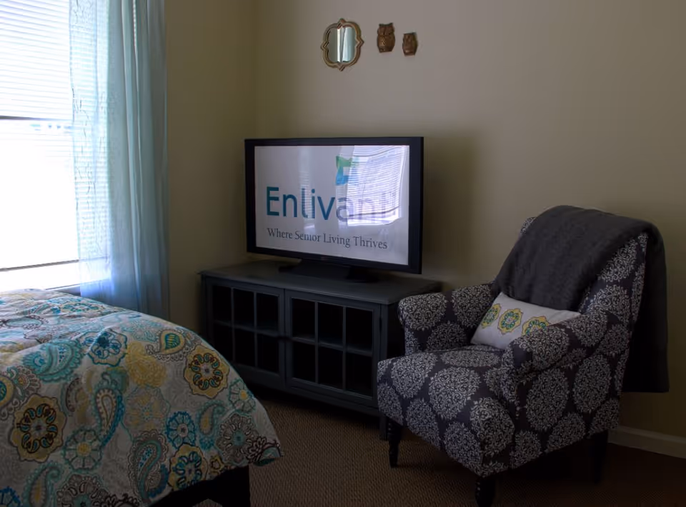 A bedroom corner with a patterned armchair, media cabinet holding a TV displaying 'Enlivant', and part of a patterned bed near a window.