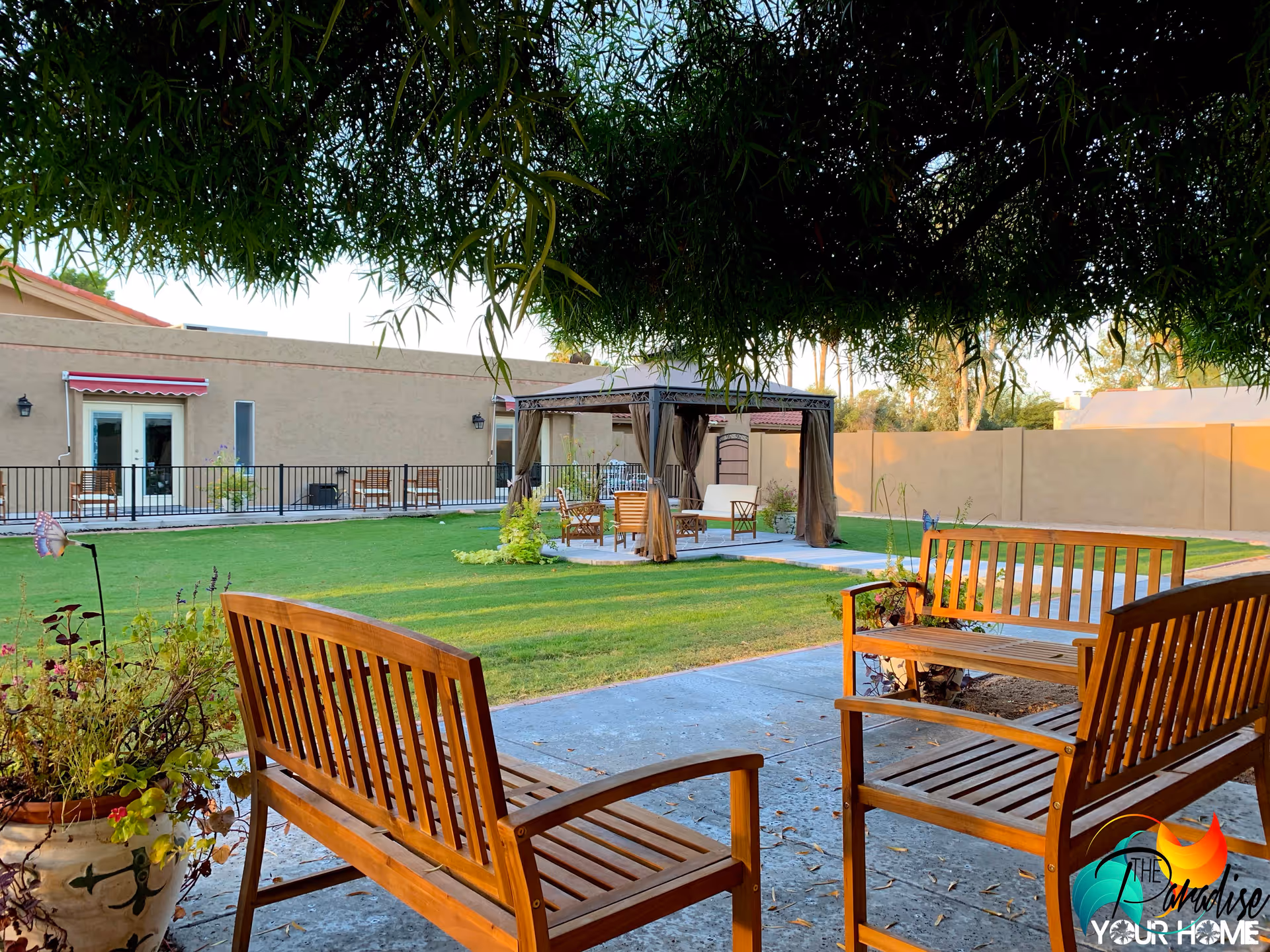 Outdoor garden area at The Paradise Assisted Living Home featuring wooden benches on a concrete patio, a grassy lawn, a gazebo with seating, and a beige building with doors and windows in the background, shaded by tree branches overhead.