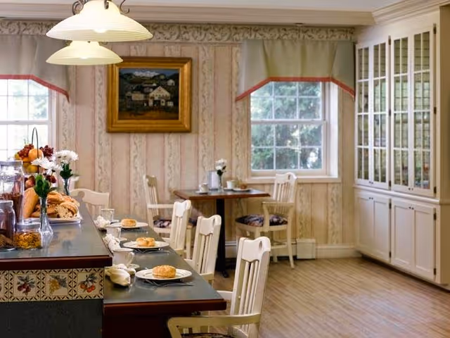 A cozy dining area in a senior living facility with a counter set for breakfast, including plates with bagels, cups, and napkins. The room features white wooden chairs, a small table with chairs near the window, floral wallpaper, a painting on the wall, and a large cabinet with glass doors.