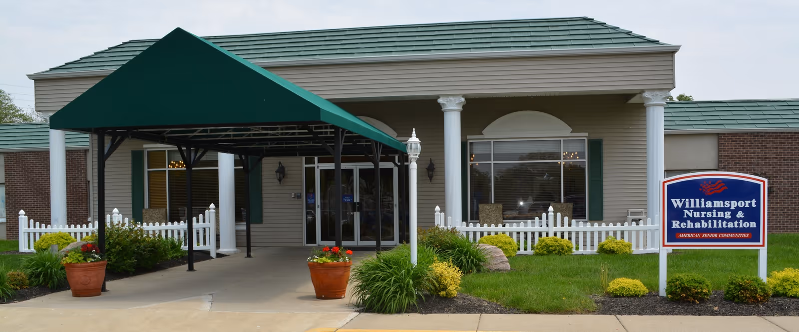 Entrance of Williamsport Nursing and Rehabilitation facility with a green canopy over the driveway, white columns, a white picket fence, and a sign displaying the facility's name on a landscaped lawn.