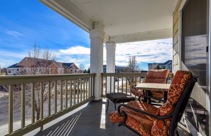 Covered balcony with patterned cushioned chairs and a small table overlooking neighborhood homes under a blue sky.