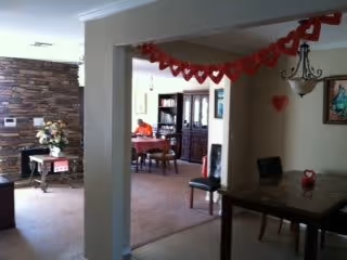 Interior view of a senior living facility showing a dining area with tables and chairs, a stone accent wall with a small table and flower arrangement, and red heart-shaped decorations hanging from the ceiling.
