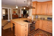 A spacious kitchen with wooden cabinets and a large island featuring a built-in stove. The kitchen has a hardwood floor and a decorative rug. Pendant lights hang above the island. In the background, there is a view into a living room area with beige furniture and a piano.