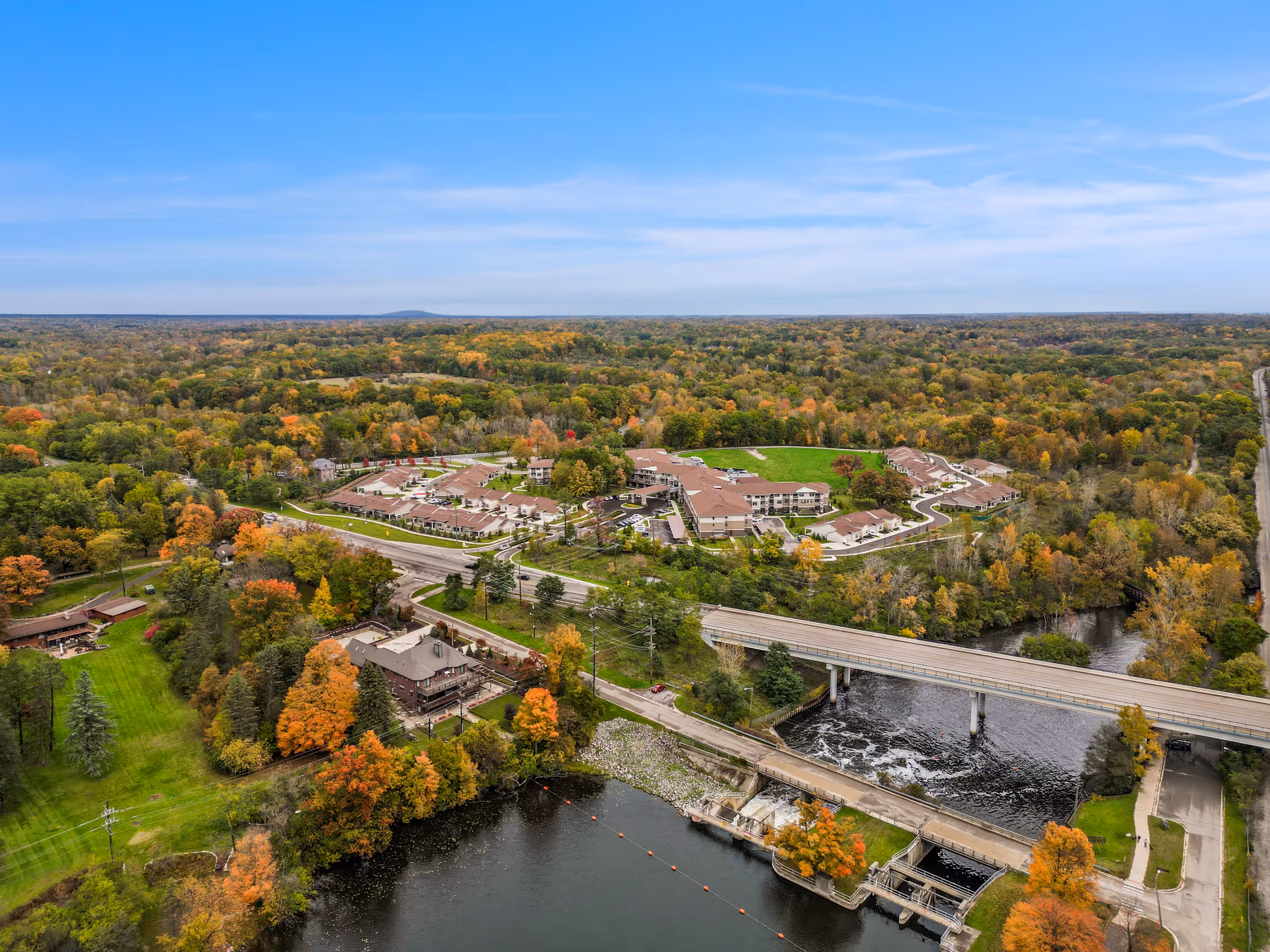 Aerial view of a residential complex surrounded by autumn trees with a river and bridge in the foreground.