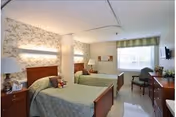 A nursing facility room with two single beds separated by a curtain. Each bed has a green bedspread and a wooden headboard. There is a bedside table with a lamp and a teddy bear on one bed. The room has floral wallpaper, a window with a green valance, a small table with two chairs, a wall-mounted television, and a wooden dresser.