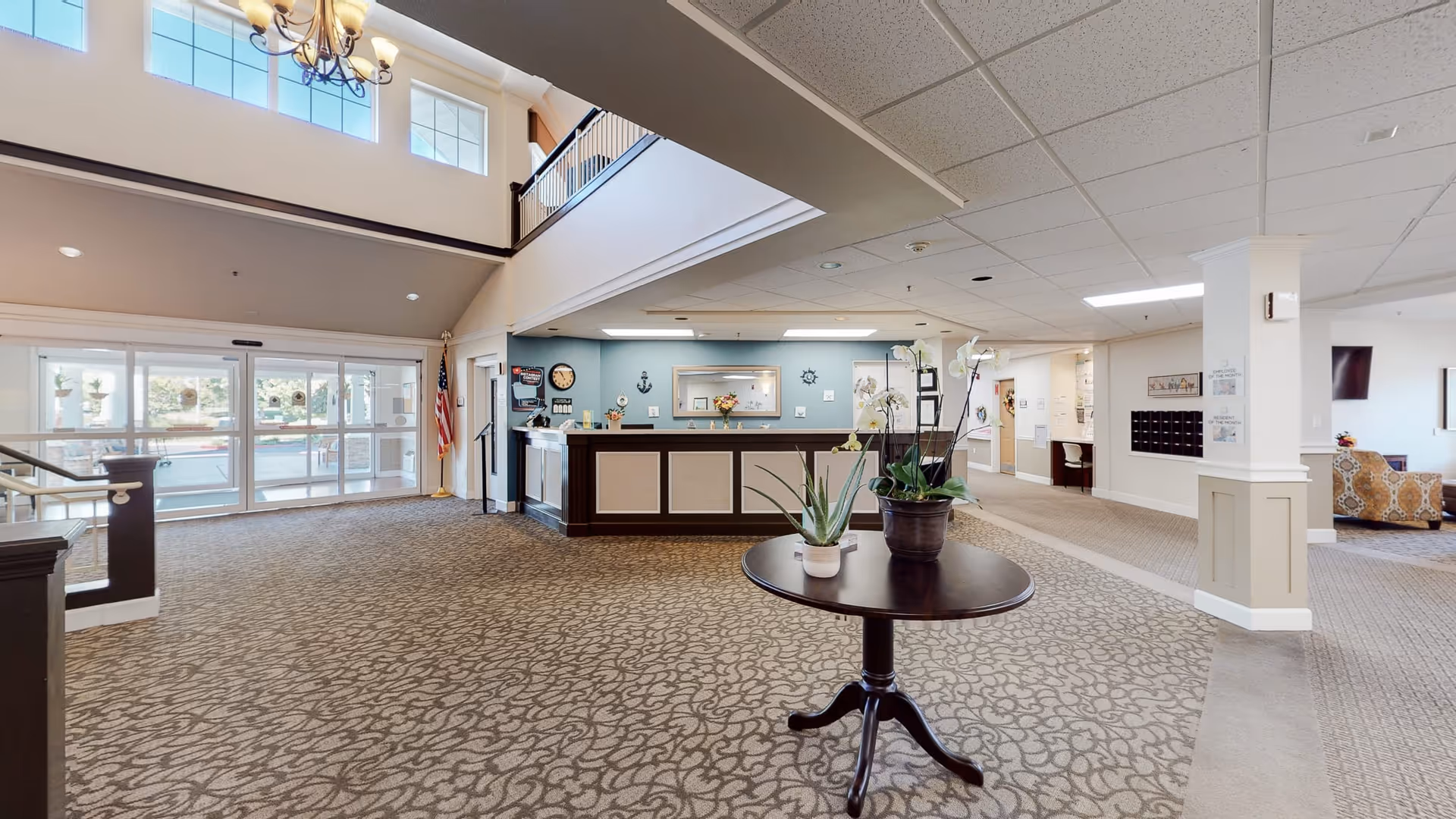 Spacious lobby area of The Commons At Dallas Ranch with a patterned carpet, a round wooden table with potted plants in the center, a reception desk against a blue accent wall, large windows and glass doors letting in natural light, an American flag near the entrance, and seating areas visible in the background.