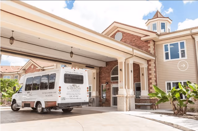 Exterior view of The Gardens at Brook Ridge Assisted Living & Memory Care entrance with a covered drop-off area and a white shuttle van parked nearby. The building features brick and beige siding with large windows and a small tower structure on the roof.