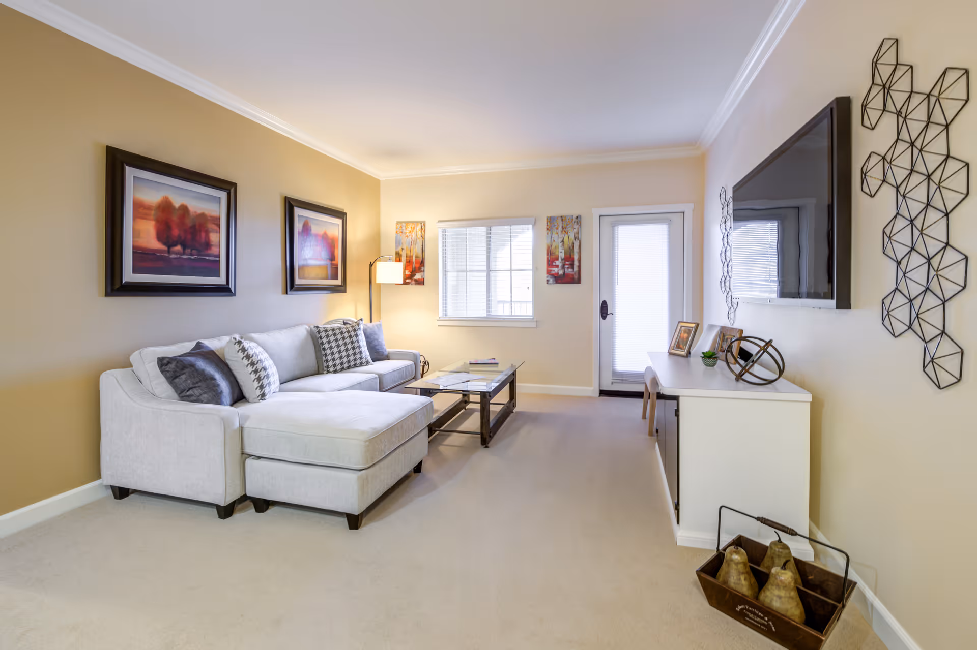 A bright living room with a light gray sectional sofa adorned with patterned pillows, a glass coffee table, and a white media console with decorative items. The walls are painted beige and white, featuring framed artwork and geometric metal wall decor. A window and a door with blinds allow natural light into the room.