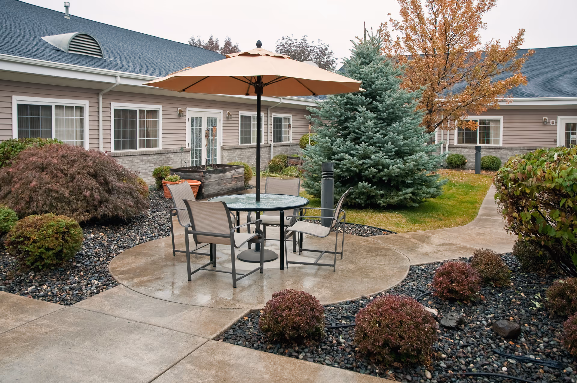 Outdoor patio area at a senior living facility with a round glass table, four chairs, and a large beige umbrella. The patio is surrounded by landscaped bushes, a small evergreen tree, and a deciduous tree with autumn leaves. The building with multiple windows and doors is visible in the background.