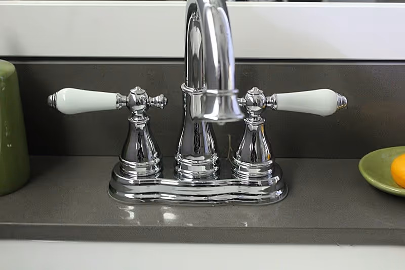 Close-up view of a chrome faucet with white handles mounted on a dark countertop, with a green container on the left and a green plate with an orange on the right.