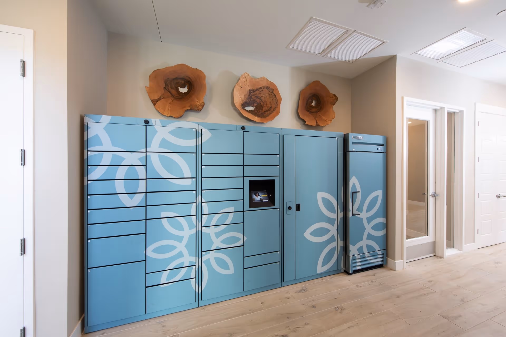 A modern indoor package locker system with multiple compartments in light blue color featuring white floral patterns. Above the lockers, there are three decorative wooden wall art pieces resembling cross-sections of tree trunks. The room has light-colored wooden flooring and beige walls, with white doors and vents on the ceiling.