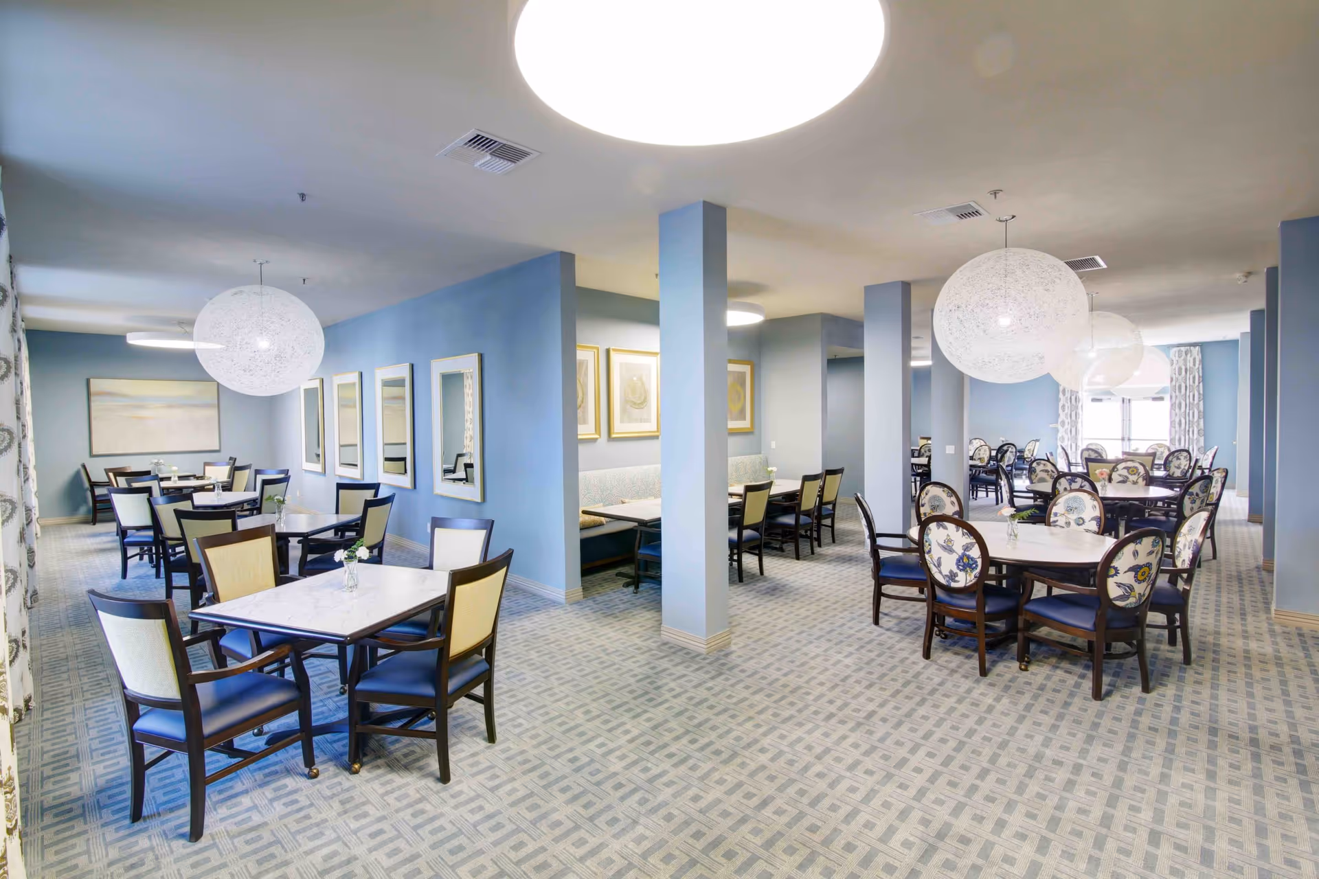 A spacious dining room in a senior living facility with multiple tables and chairs arranged neatly. The walls are painted light blue and decorated with framed artwork and mirrors. Large spherical pendant lights hang from the ceiling, and the carpet has a geometric pattern. The room is well-lit with natural light coming through windows with patterned curtains.