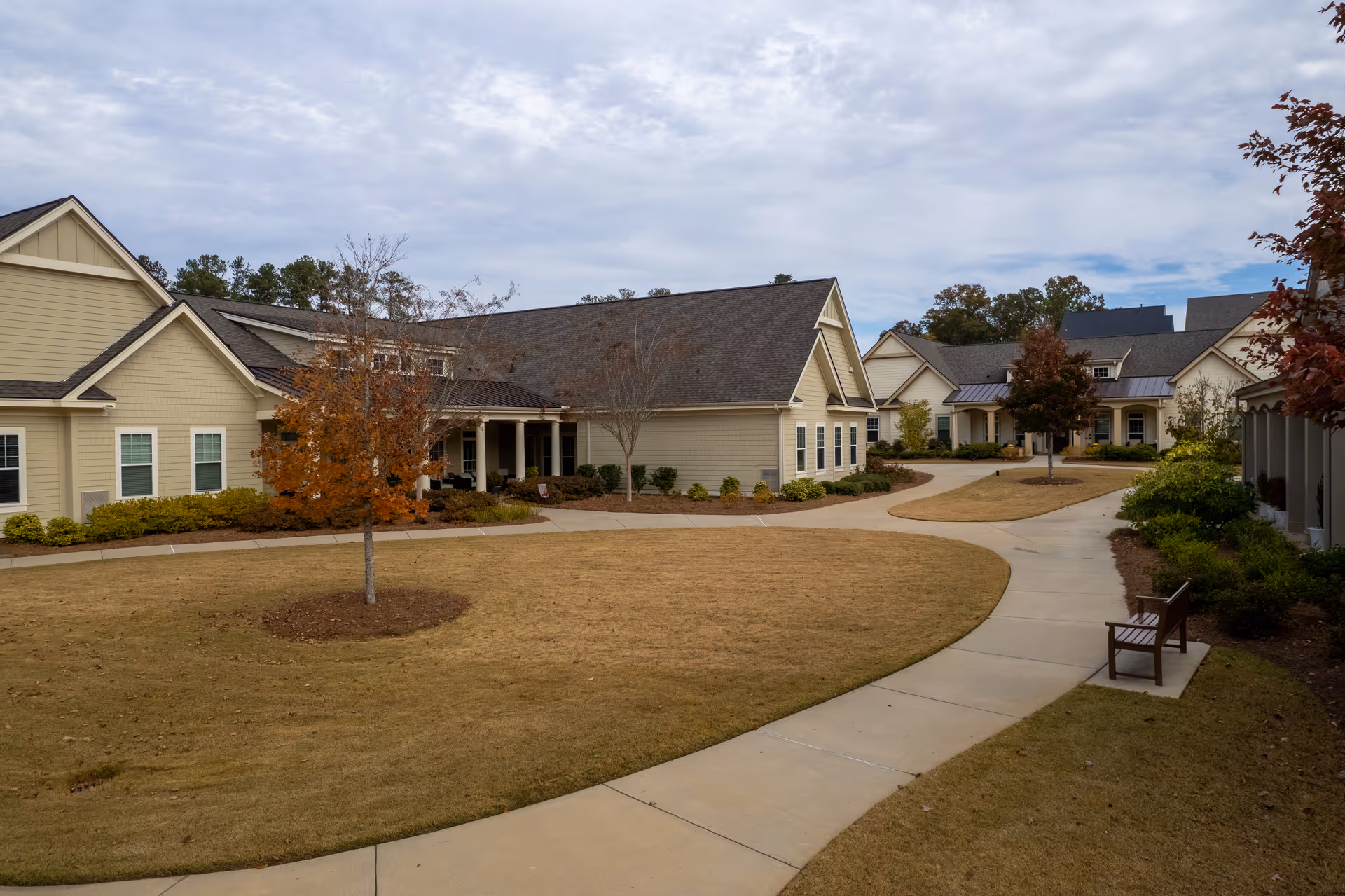 Outdoor view of a senior living community with beige buildings, a curved concrete walkway, a few small trees with autumn leaves, and a wooden bench on the right side. The sky is partly cloudy.
