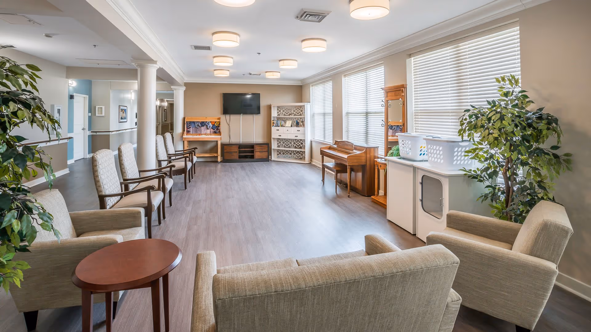 A spacious and well-lit common area in a senior living facility with multiple chairs arranged along the walls and around small tables. The room features large windows with blinds, a piano, a television mounted on the wall, and some plants adding greenery to the space. The flooring is wood-style, and the ceiling has multiple round light fixtures.