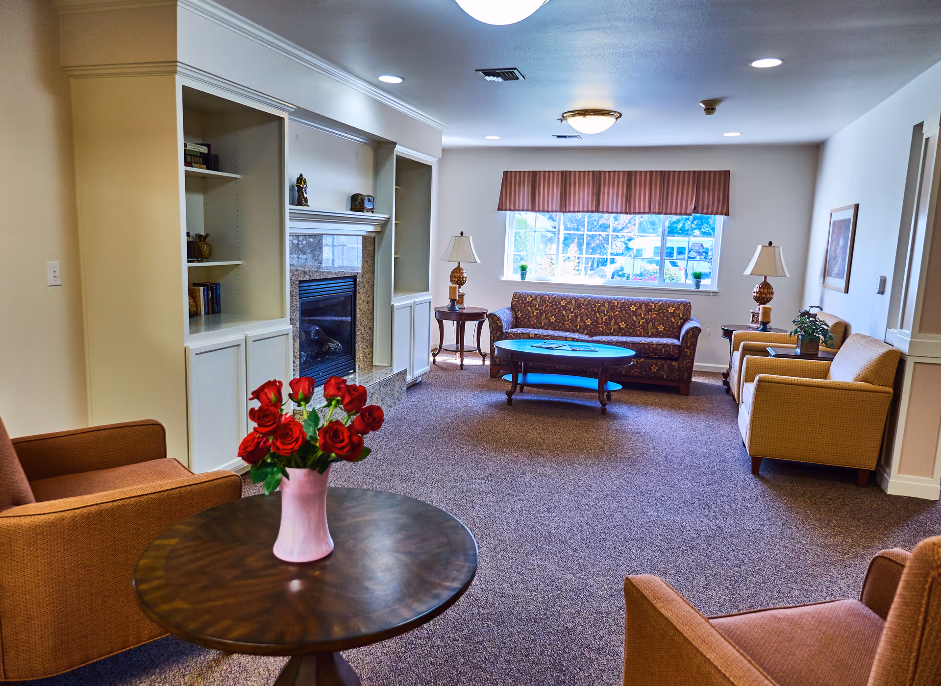 A cozy living room in South Pointe Assisted Living featuring a floral-patterned sofa, two armchairs, a round wooden coffee table with a vase of red roses, side tables with lamps, a fireplace with built-in shelves, and a large window with a valance letting in natural light.