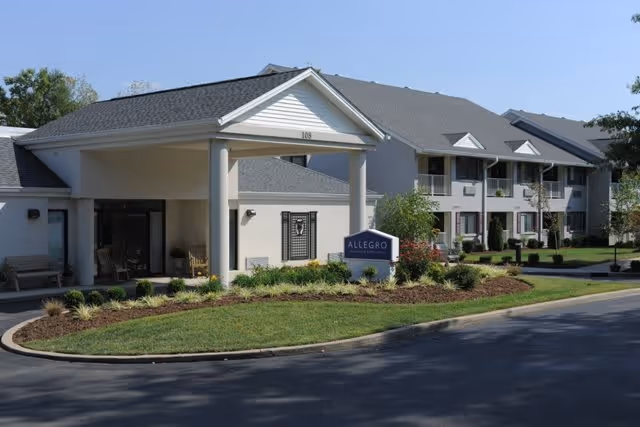 Exterior view of a senior living facility building with a covered entrance, well-maintained landscaping, and a sign that reads 'Allegro'. The building has two stories with balconies and multiple windows under a clear blue sky.