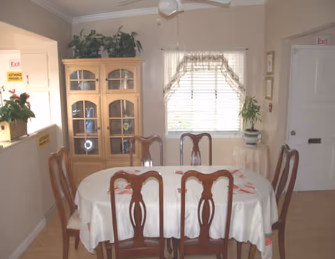 A dining room with a round table, white tablecloths, wooden chairs, cabinet, and plants.