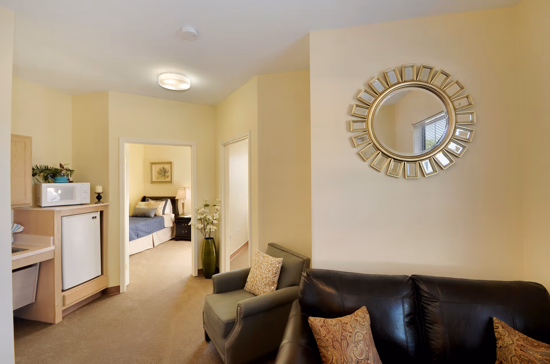 Interior view of a senior living facility room showing a small living area with a black leather sofa and a gray armchair with patterned pillows. A decorative round mirror with a gold frame hangs on the wall above the sofa. In the background, there is a small kitchenette area with a microwave and mini fridge, and an open doorway leading to a bedroom with a bed, nightstand, lamp, and framed artwork on the wall.