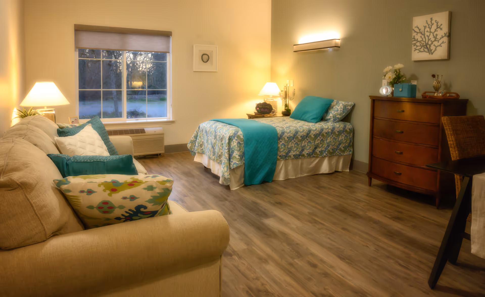 A cozy bedroom in a senior living facility featuring a single bed with blue and patterned bedding, a beige sofa with colorful pillows, a wooden dresser with decorative items, a window with a view of trees outside, and warm lighting from lamps.