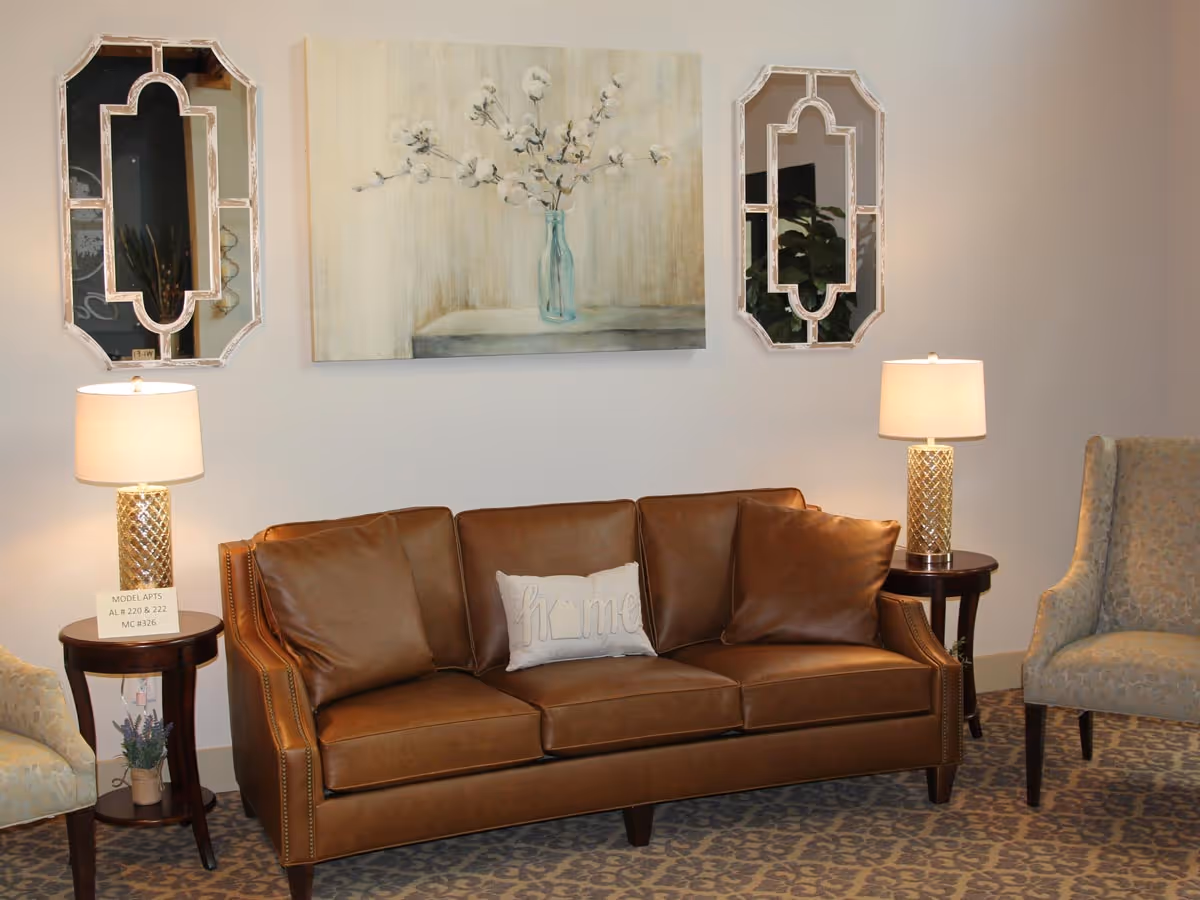 Cozy seating area with a brown leather sofa flanked by two lamps, mirrors, artwork, and upholstered chairs.