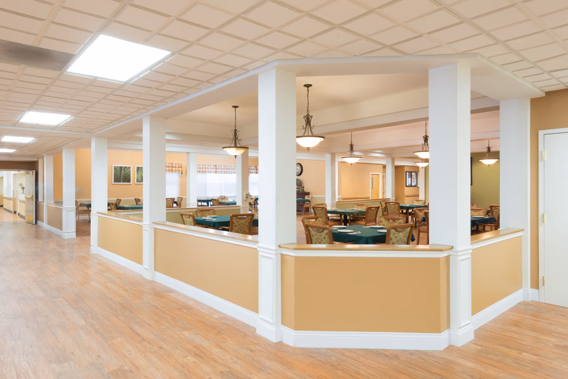 Interior view of a senior living facility dining room with multiple round tables covered with green tablecloths and surrounded by chairs. The room is well-lit with hanging light fixtures and has beige walls with white trim. The floor is wooden, and there is a hallway visible to the left.