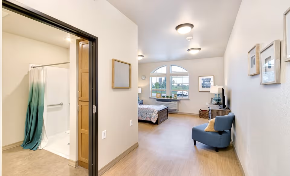 A bright and clean senior living bedroom with a large window, a bed with patterned bedding, a blue armchair with a pillow, a wooden dresser with a TV, and framed artwork on the walls. To the left, there is an open doorway leading to a bathroom with a shower and a green and white shower curtain.
