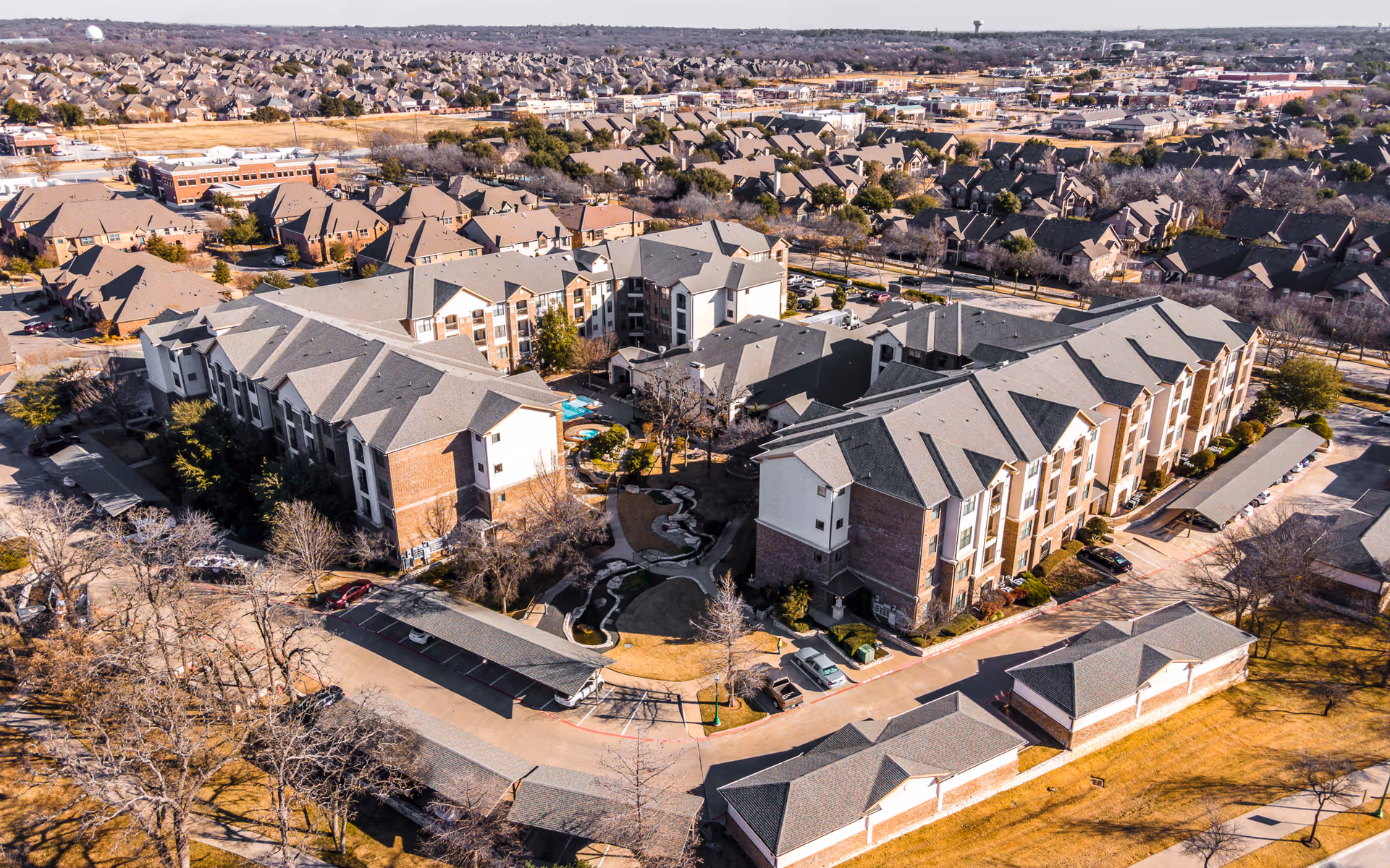 Aerial view of Conservatory At Keller Town Center, showing multiple connected residential buildings with gray roofs and brick facades, surrounded by parking areas, walkways, and landscaped grounds. The surrounding neighborhood with many houses is visible in the background.