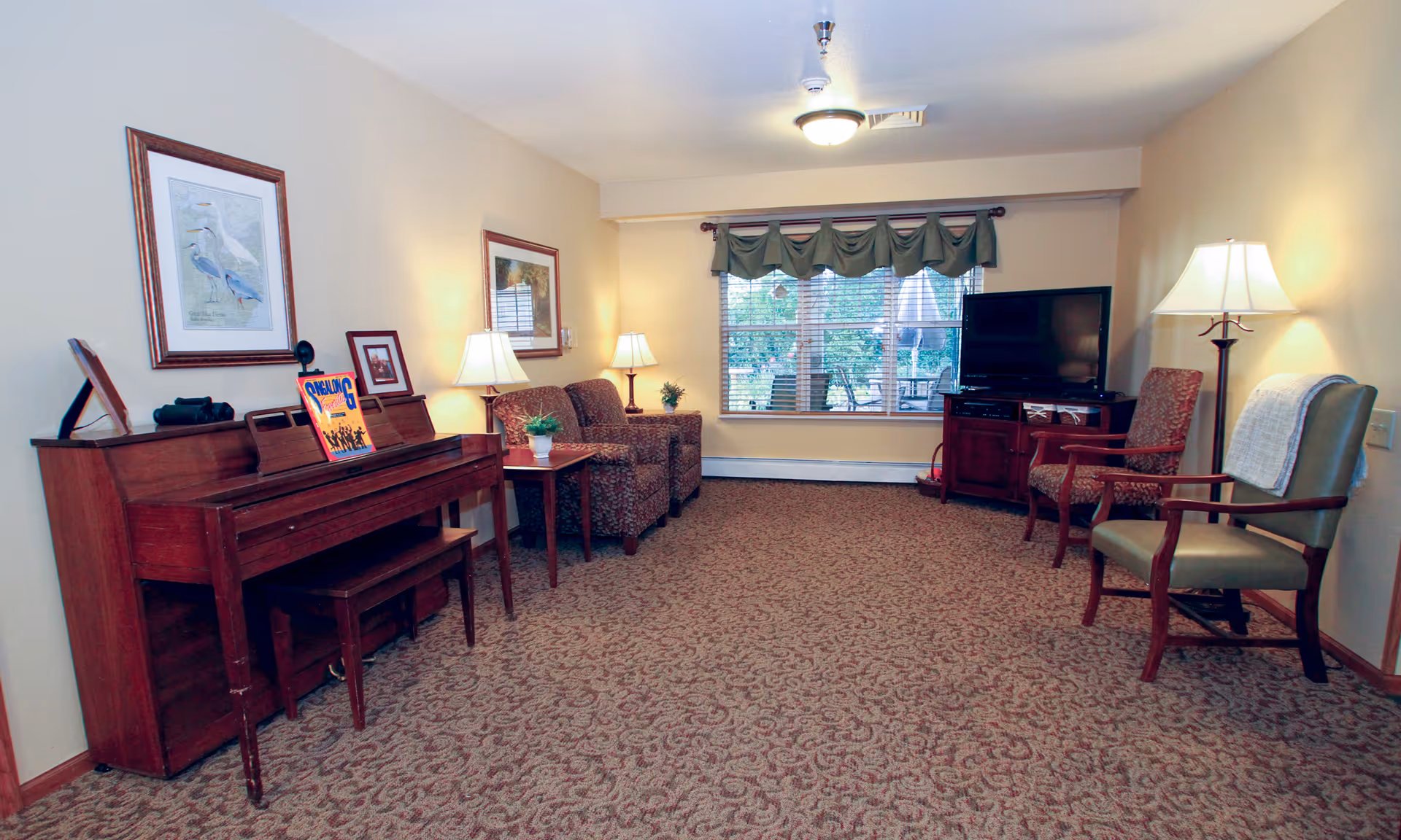 A cozy living room with patterned carpet, two upholstered armchairs, a wooden piano with a bench, framed artwork on the walls, two table lamps, a floor lamp, a flat-screen TV on a wooden stand, and a large window with green valance curtains showing an outdoor patio area.